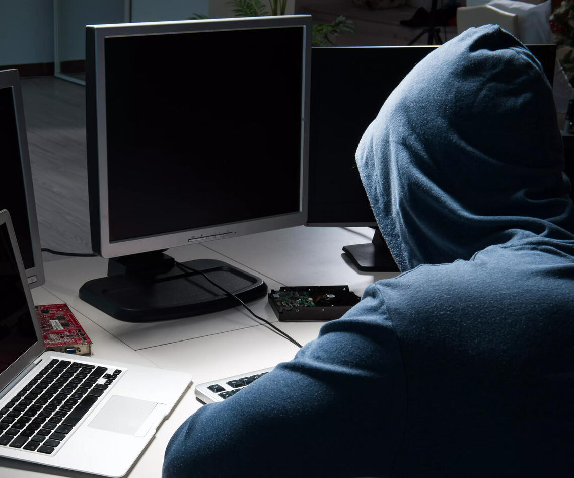 A hooded figure working on multiple computer screens