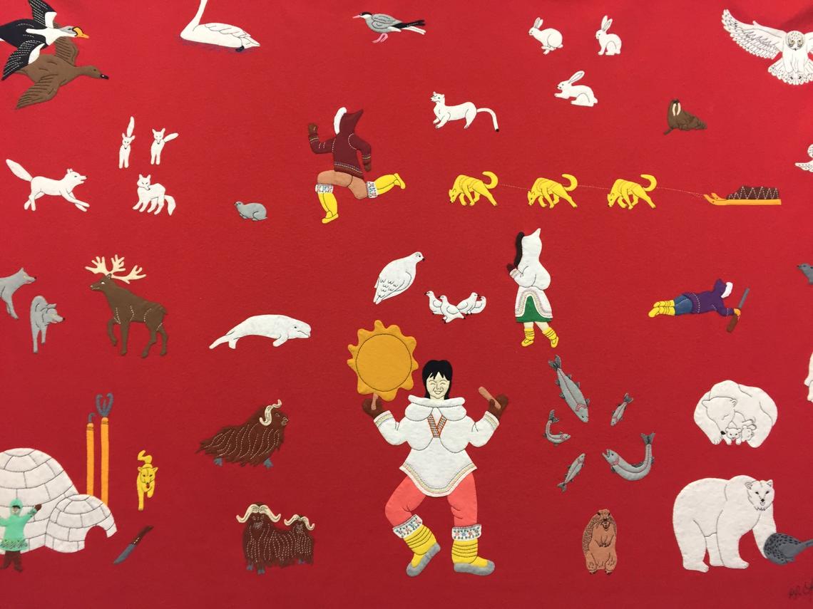 Image is of an Inuit wall hanging, made of felt. The background is bright red, and stitched on the fabric are felt-stitched images of a drum dancer, an iglu, Arctic animals including the polar bear, seals, whale.