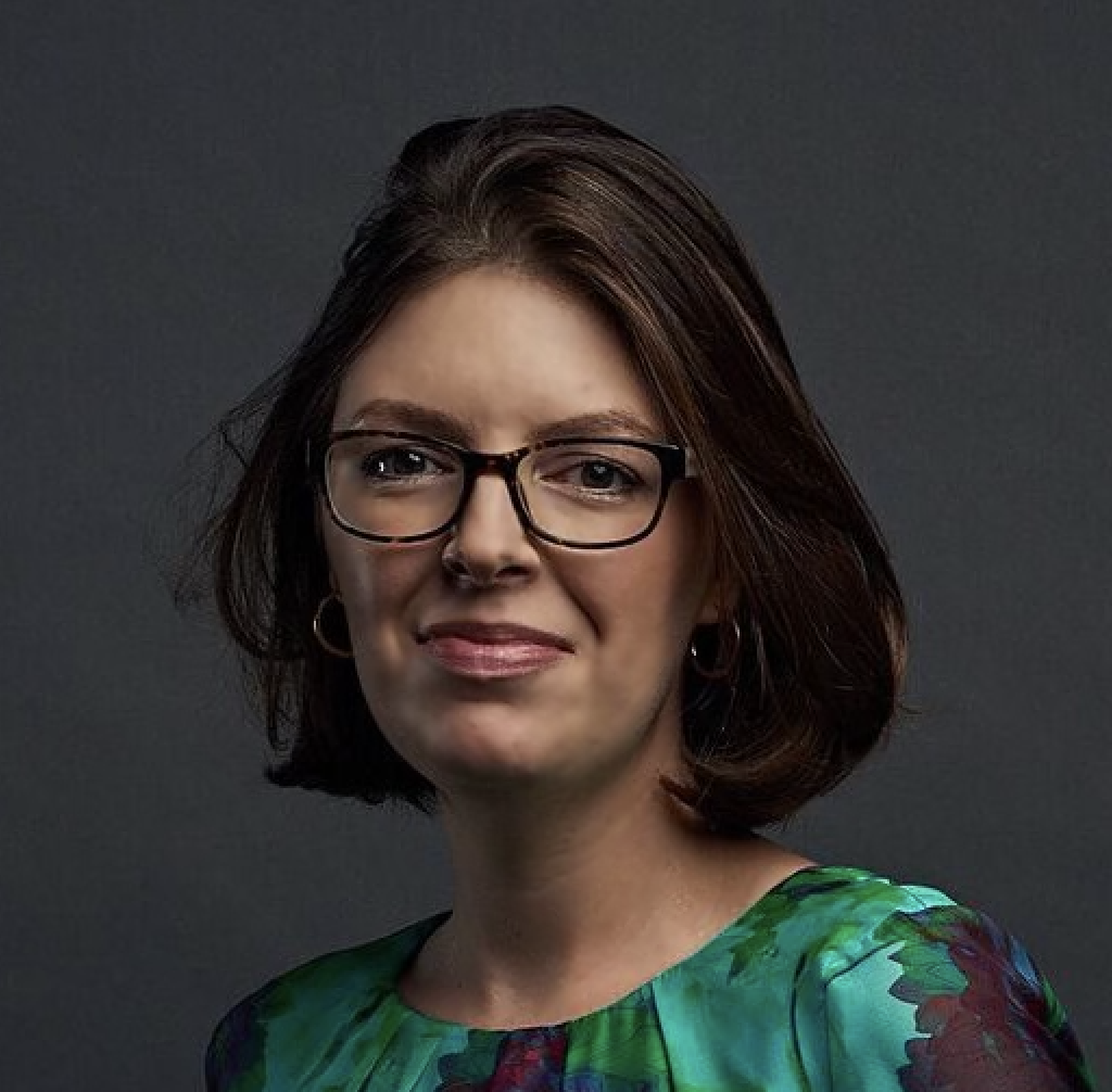 Myka has brown hair and glasses and is wearing a green shirt. She is smiling at the camera.