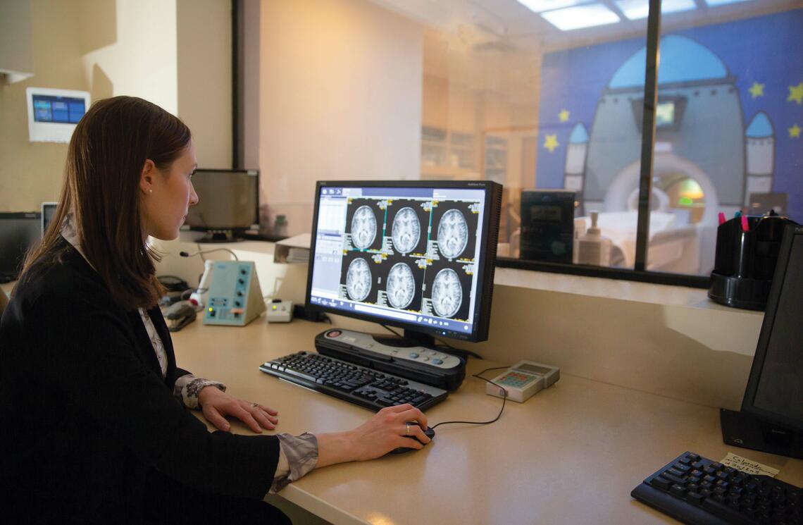 A photograph of a lady sitting in front of a computer screen looking at brain scans 