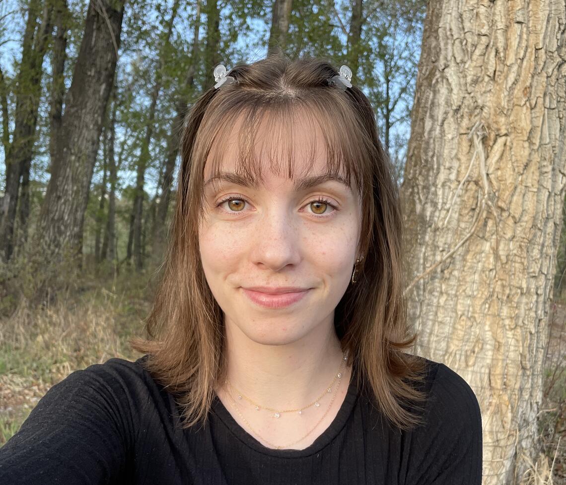 Maggie has brown hair and pale skin. She is standing in a forest.