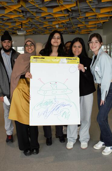 A group of students pose around a posterboard with a drawing on it. 