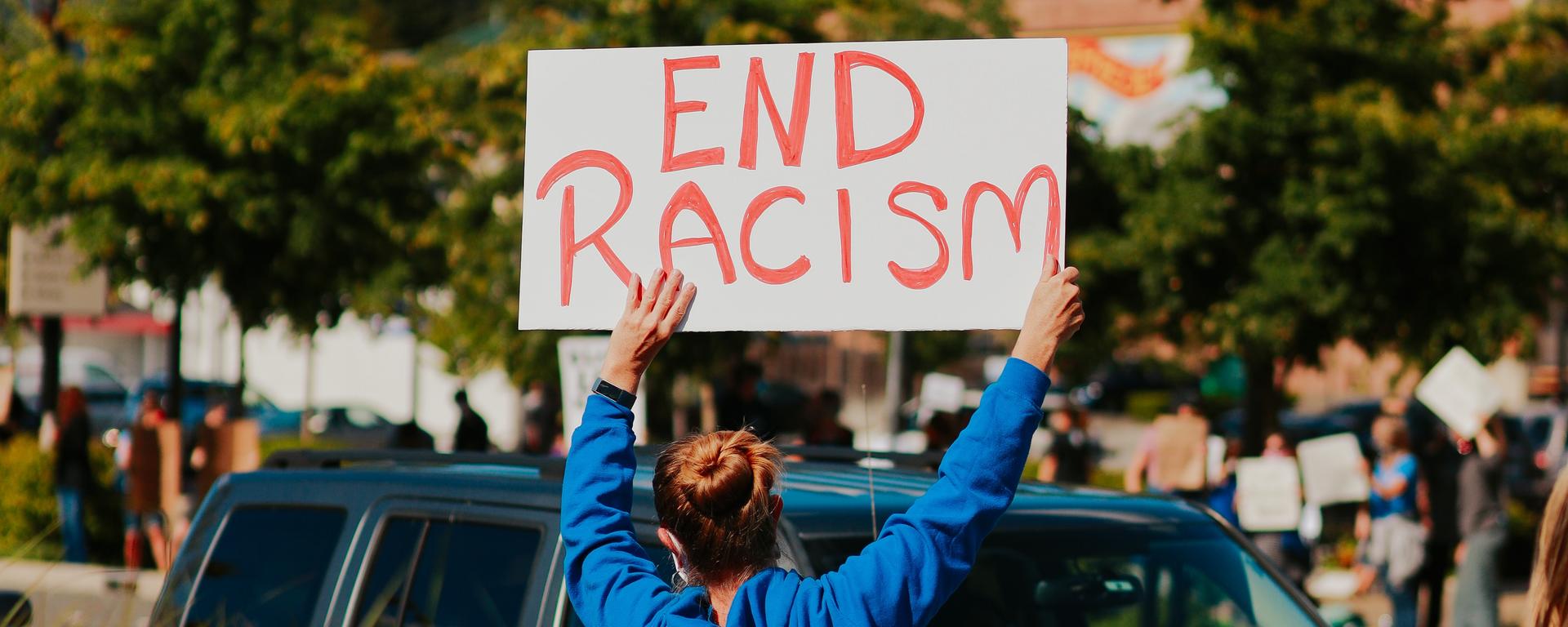 A woman faces traffic and holds up a sign that says "End Racism"