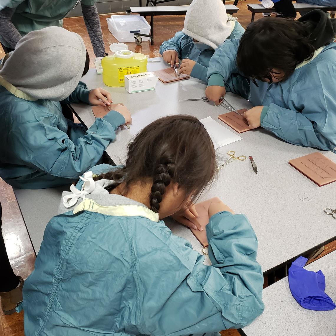 A group of children in blue scrubs practise stitching on a suture pad. 