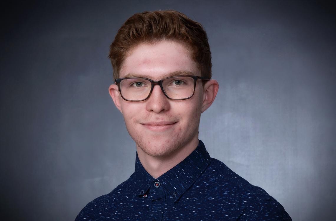 Young man with dark red hair wearing a navy button up shirt with white flecks and dark tortoiseshell glasses