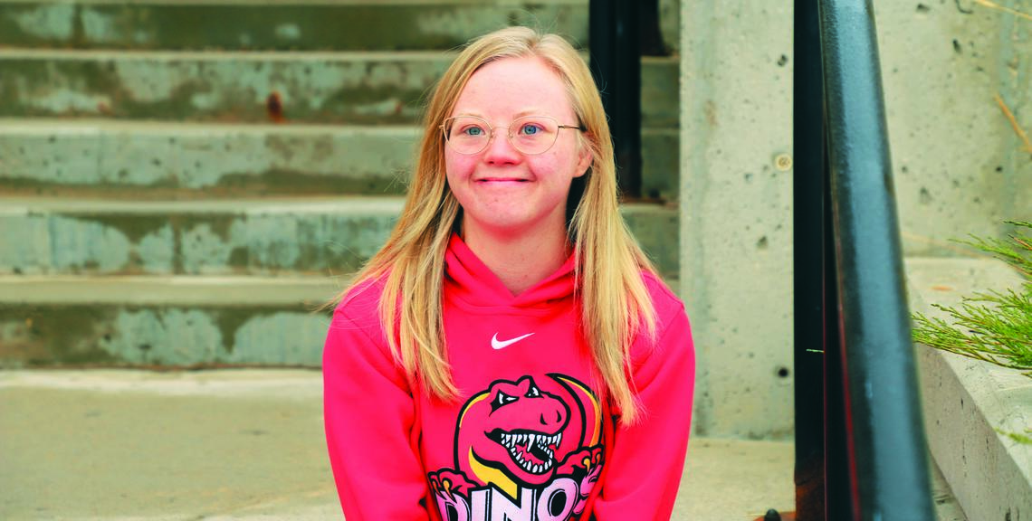 Young blonde woman wearing a red hoodie with the Dinos logo and gold glasses, sitting on stairs