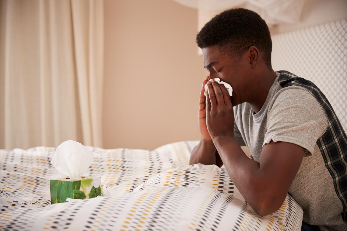 A man sitting in bed and blowing his nose with a tissue.