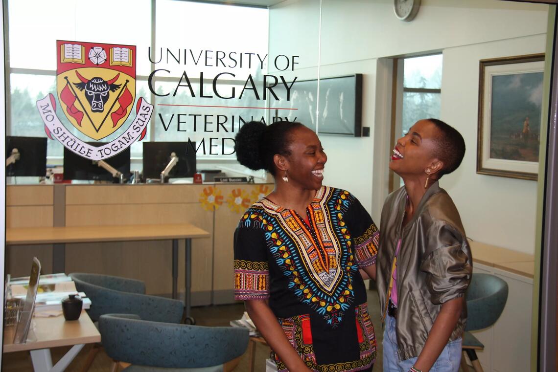 Two people stand in front of a University of Calgary sign smiling and looking at one another