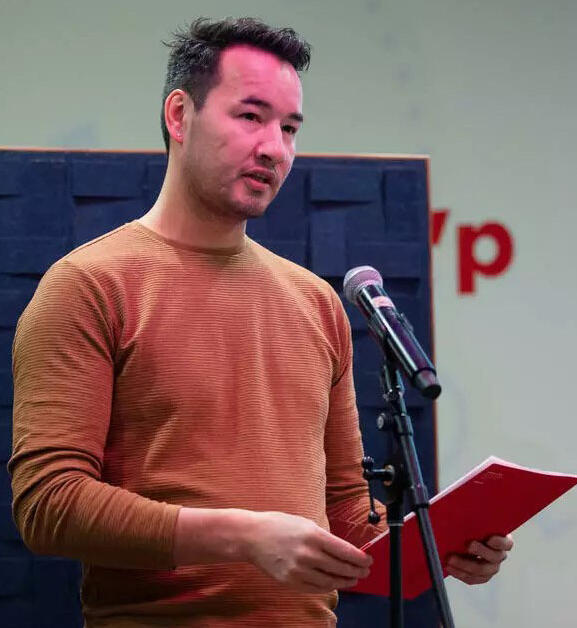 A man in a sweater stands in front of a microphone holding a red paper