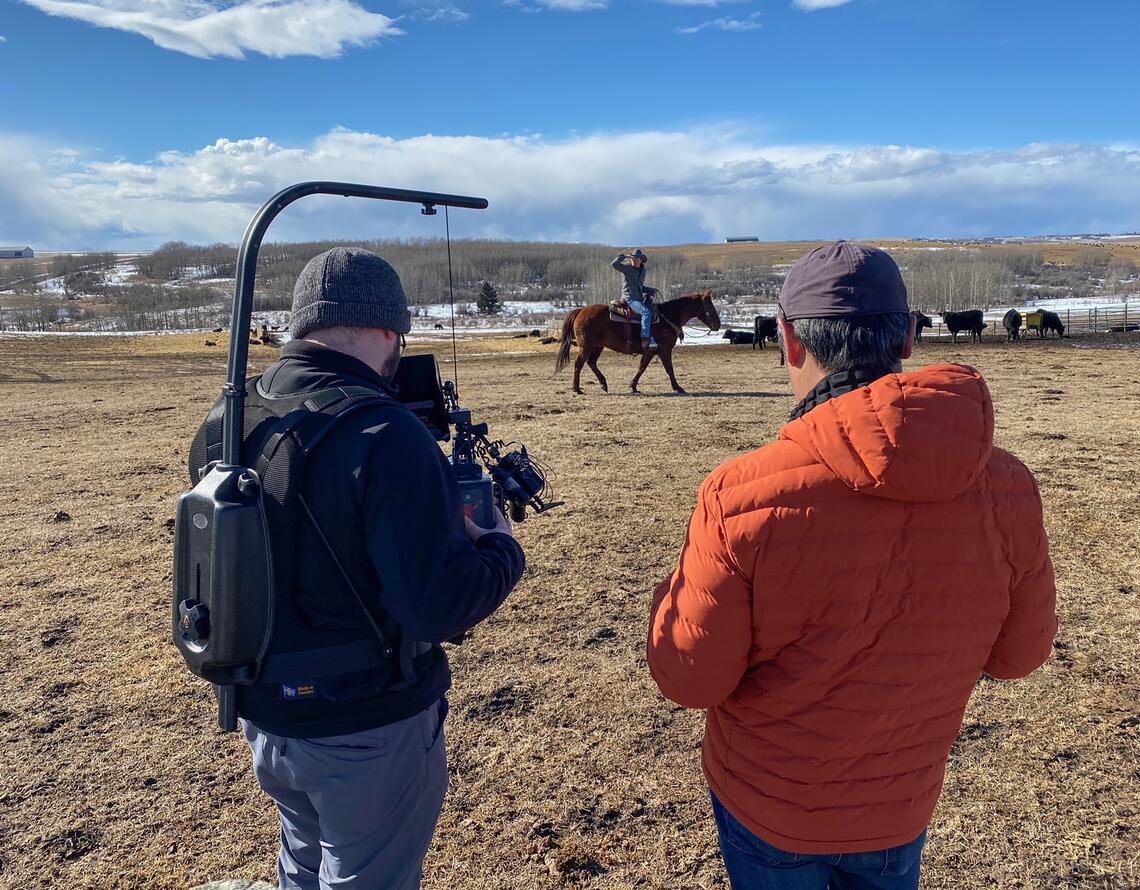 A group of two film a man riding a horse in a field on a ranch