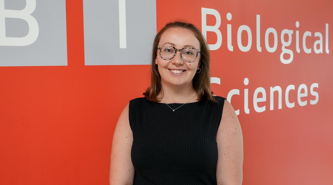 A woman with short brown hair and glasses stands in front of a red wall