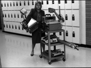 A communications media assistant carries an overhead projector while pushing a cart laden with equipment that includes two slide projectors, a film projector and an overhead projector. 1981.