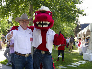 President Ed McCauley wears a white collared shirt and cowboy hat and poses in front of a tree with the UCalgary dino mascot Rex, who wears a white jean vest and cowboy hat.