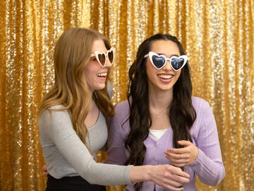 Two women wearing sunglasses smile in front of a gold background