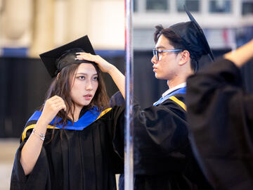 Two people in graduation regalia adjust their caps in a mirror