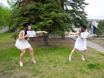 Two graduates holding their degrees and pointing at eachother