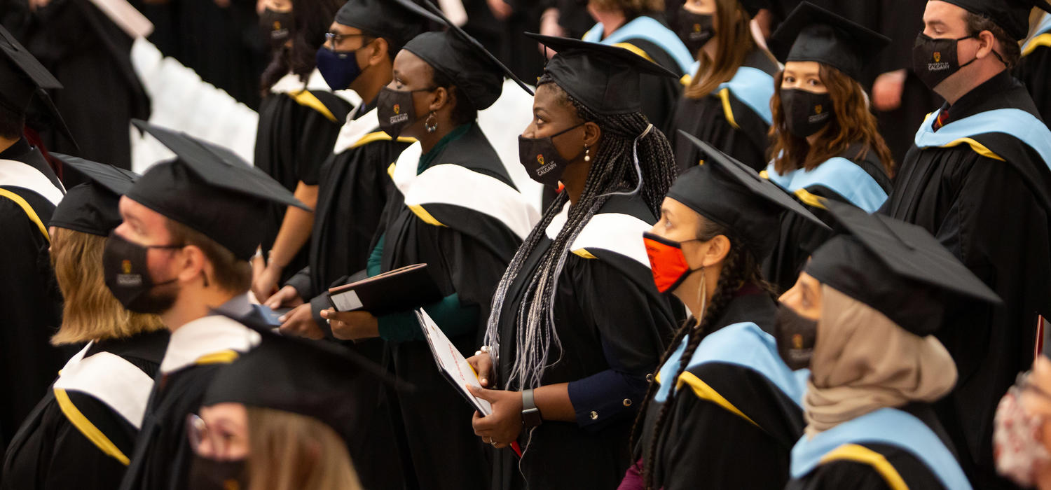 Graduates stand in their black caps and gowns, holding their degrees as they watch the stage during a convocation ceremony in 2021.. 