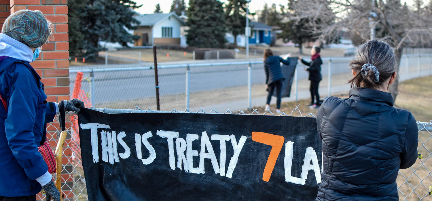 A person in a blue coat and a person in black coat hold up a black banner that says "this is Treaty 7 land" in white and orange letters.