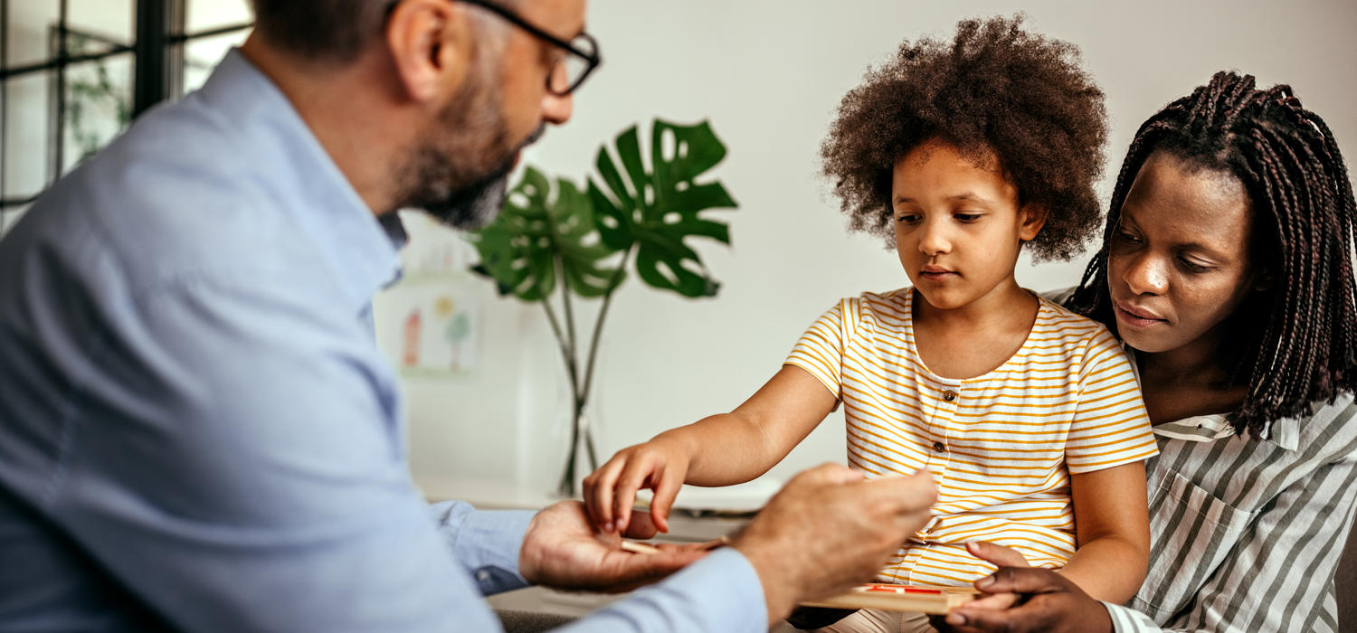 Male healthcare practitioner sitting in a room with a Black mother and her child, interacting with a puzzle.
