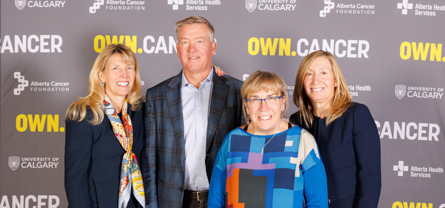 4 people stand next to each other smiling at the camera in from of a banner that has University of Calgary, Alberta Cancer Foundation, Alberta Health Services and OWN.CANCER written on it