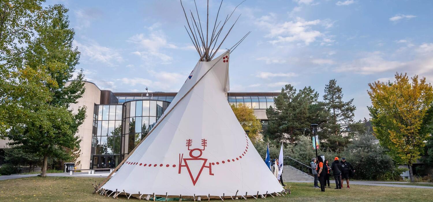 On a grassy hill, a white tipi sits in front of a glass building