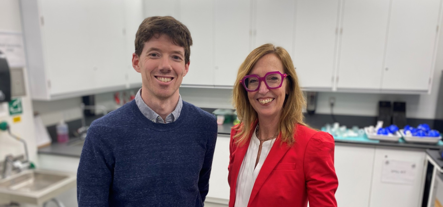 Martin MacInnis, a white man with light brown hair and wearing a blue cardigan over a collared shirt, stands beside Cari Din, a white woman with medium length blonde hair, wearing a red blazer and big red glasses. They are in a lab.