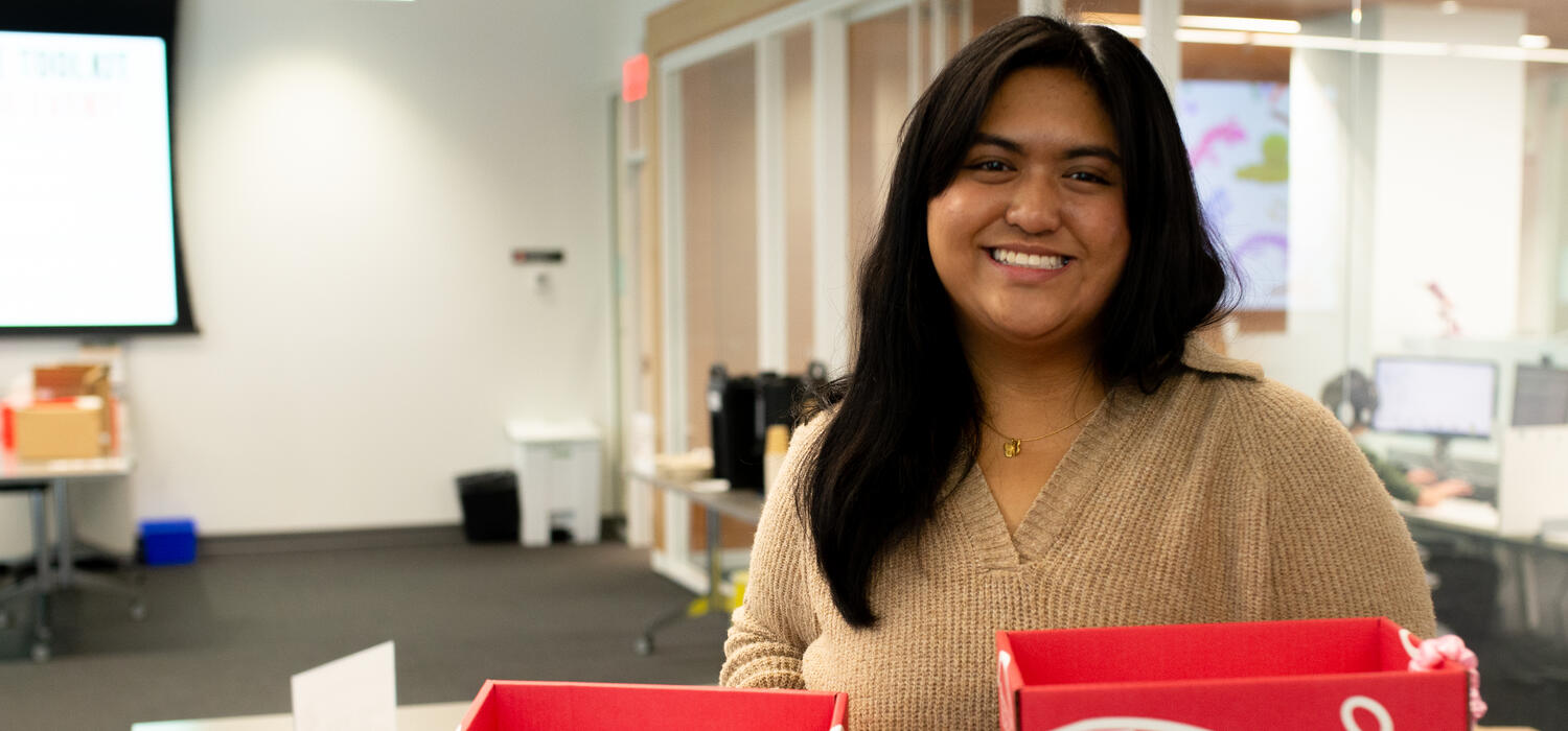 A woman stands in a classroom holding two red boxes filled with 3D printed sensory tools