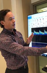 Aaron Phillips stands in front of a graph which shows how monitoring cerebral blood flow can test if enough blood is getting to the brain.