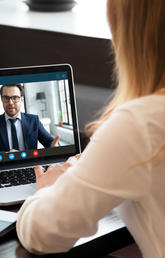 A female figure sits in front of a laptop, with a male dressed in a suit on the laptop screen.