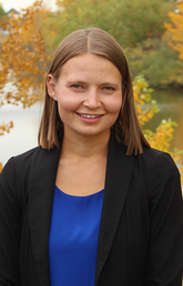 Jill de Grood wears a black blazer and blue shirt and smiles at the camera in front of an autumnal river