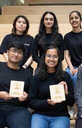 Five students and professor sitting on stairs holding championship plaques.