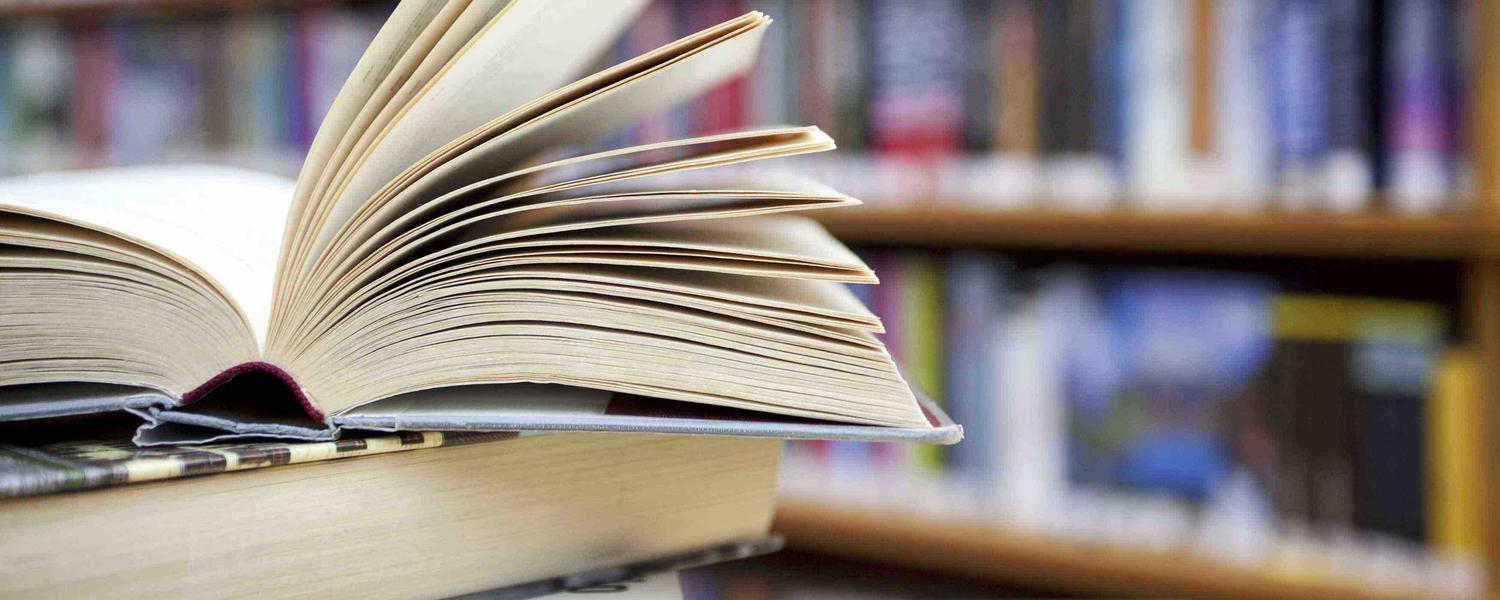 Image of a stack of books with one book opened on top and library shelves behind