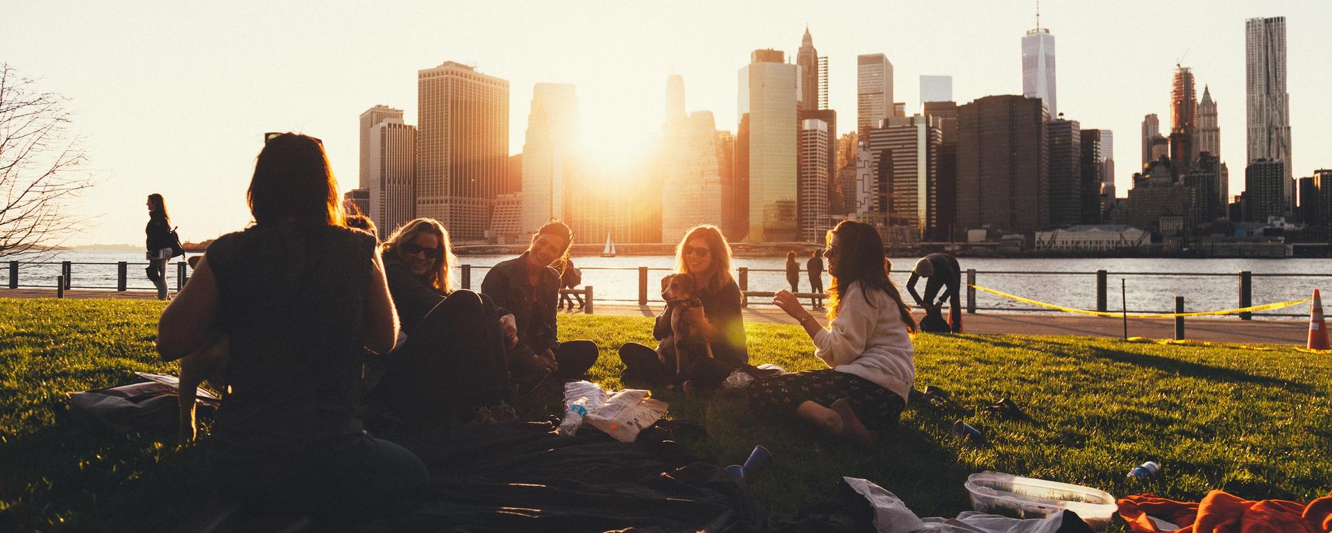 A group of students chatting, backlit by the sun, sitting in a riverside park looking towards downtown skyscrapers