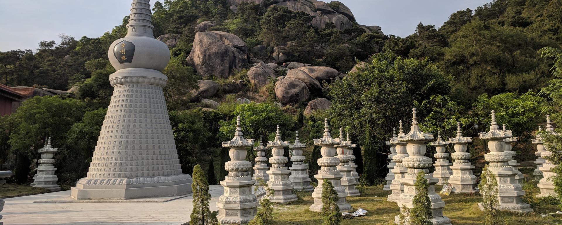 A series of stone statues (one large and white, the others small and crumbling) with a forested hillside in the background
