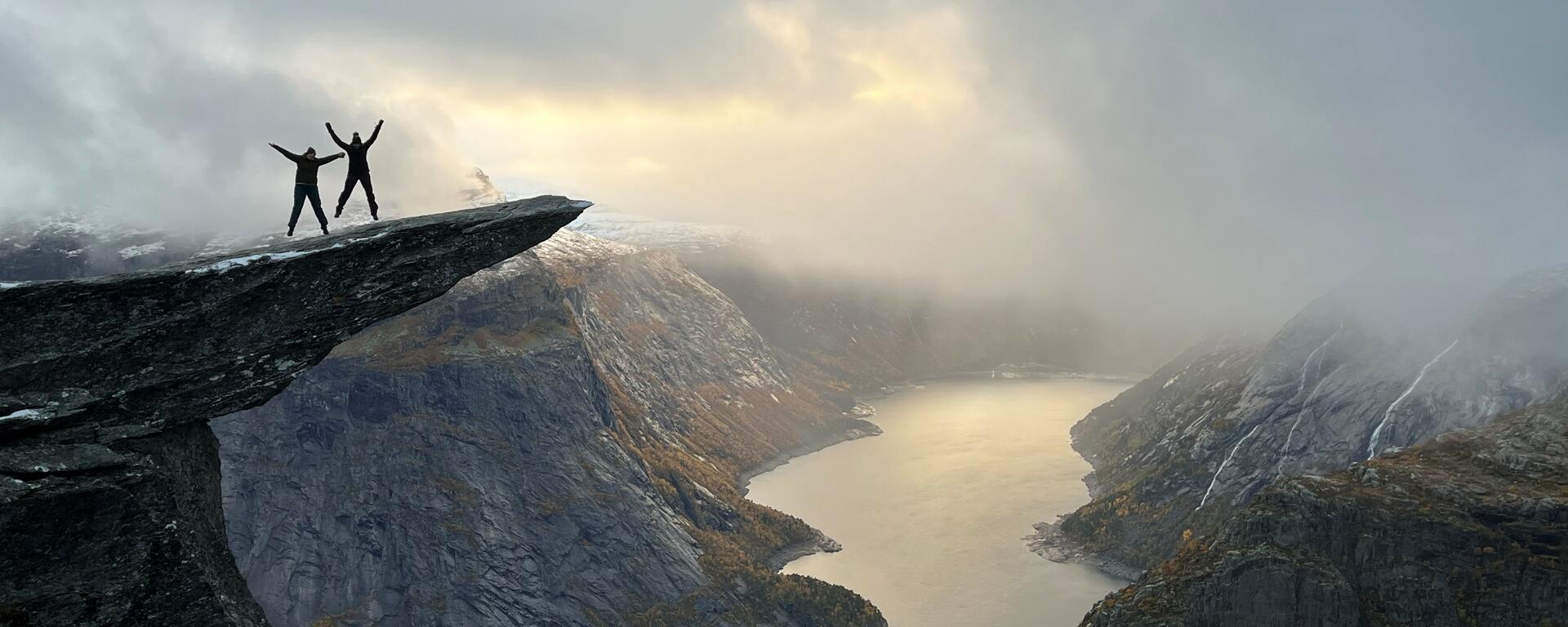 Two silhouetted figures, jumping with arms outstretched, on a narrow cliff overlooking a fjord with sun peeking through the clouds