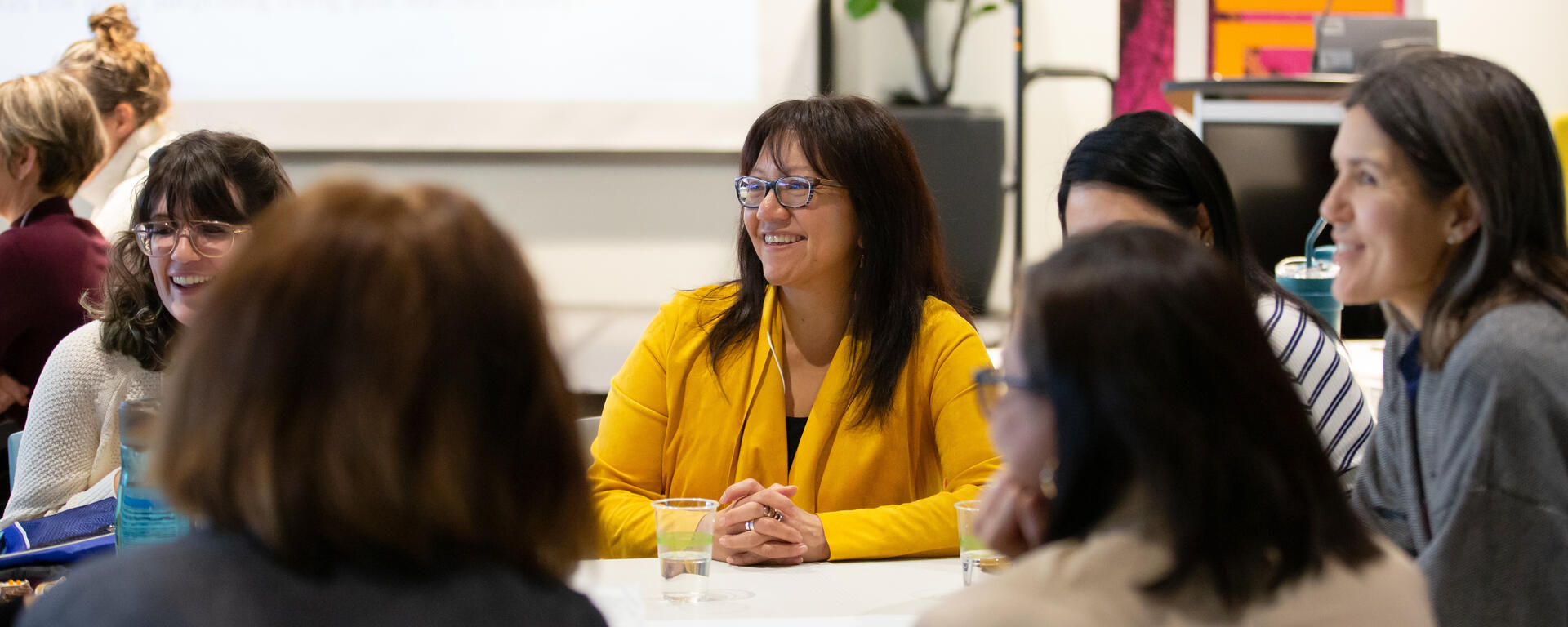 Group of people sitting at a table engaging in conversation. A woman in a yellow jacket has a smile on her face, she is the focal point of the photo.