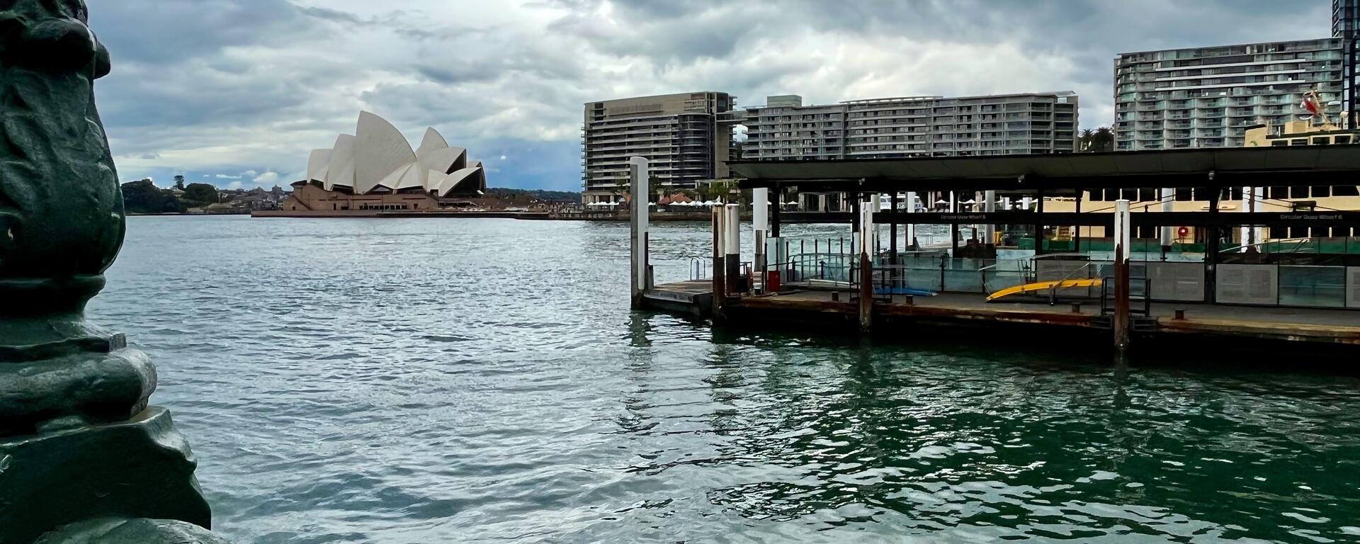 View looking across a stormy blue ocean bay. There is a wooden pier nearby on the right side. In the background, skyscrapers and the Sydney Opera House are visible along the opposite shore.
