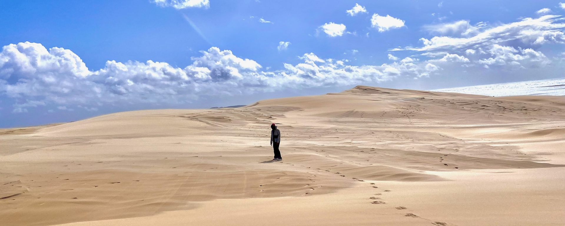 Sand dunes roll out to the horizon with blue sky above. A pair of footprints leads from the bottom right corner to the middle, where a distant silhouette of a person is walking.