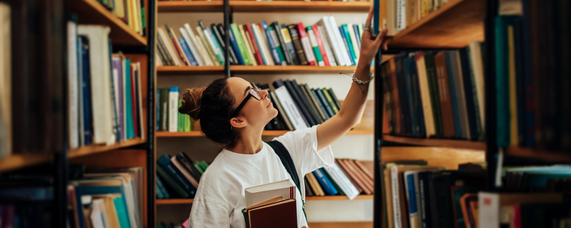 A woman with fair skin, dark hair pulled into a bun, and dark framed glasses, is holding a few books and reaching above to get a book off of a shelf in a library.