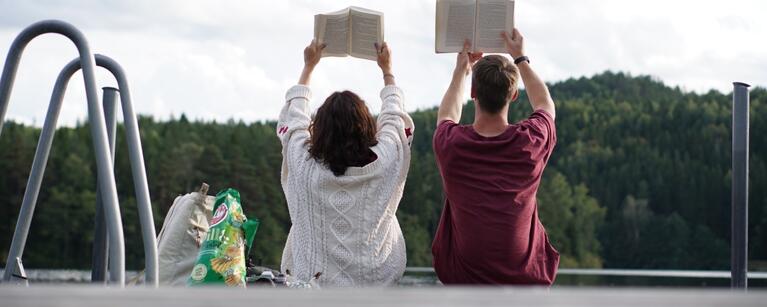 Two people, one with shoulder length brown hair and one with short cropped brown hair, sit on the edge of a dock with their backs to the camera. They both hold open books over their heads. Past the dock a strip of calm water is visible with a pine tree forest on the opposite shore.