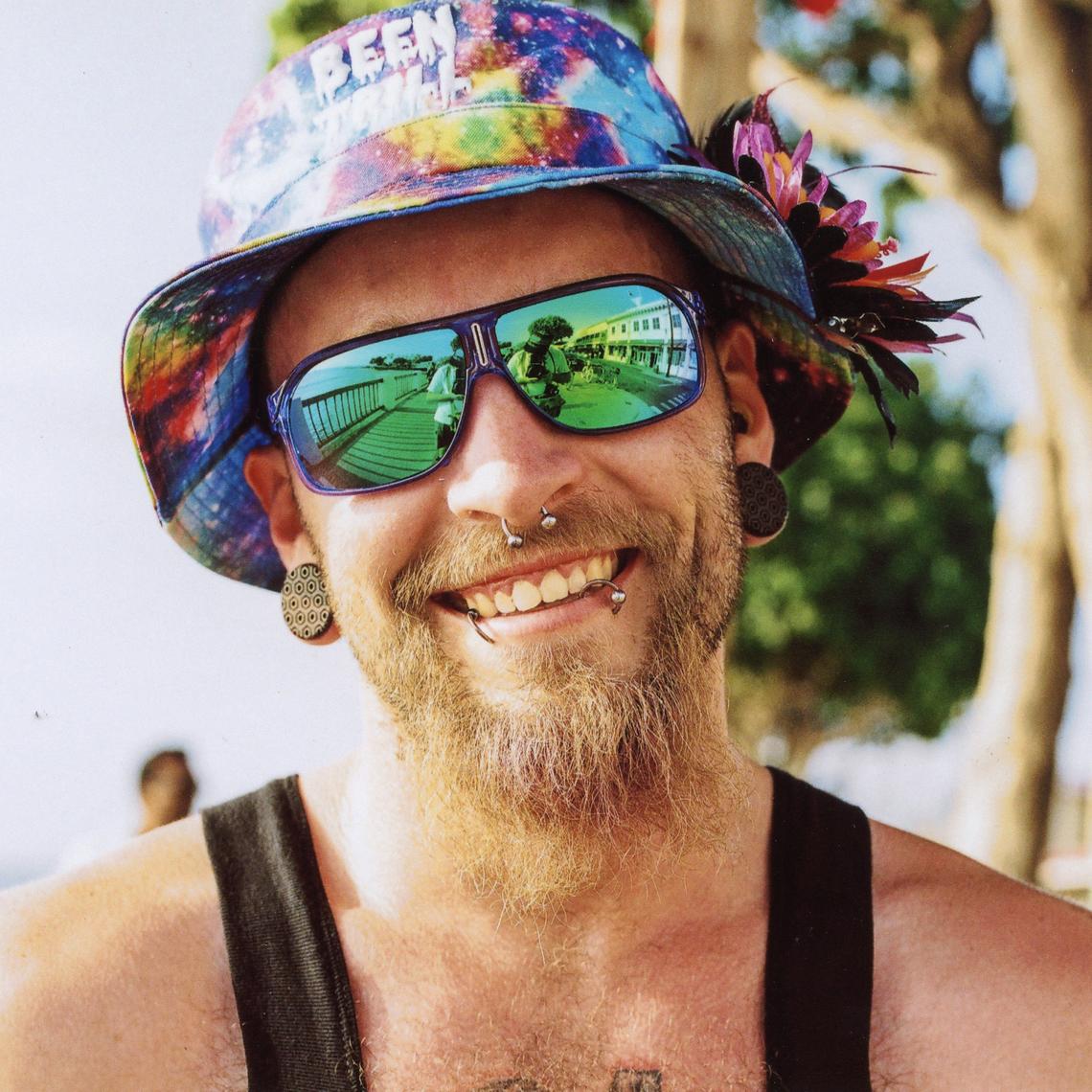 Portrait of a smiling student with a blonde beard, mirrored sunglasses, rainbow bucket hat, and piercings