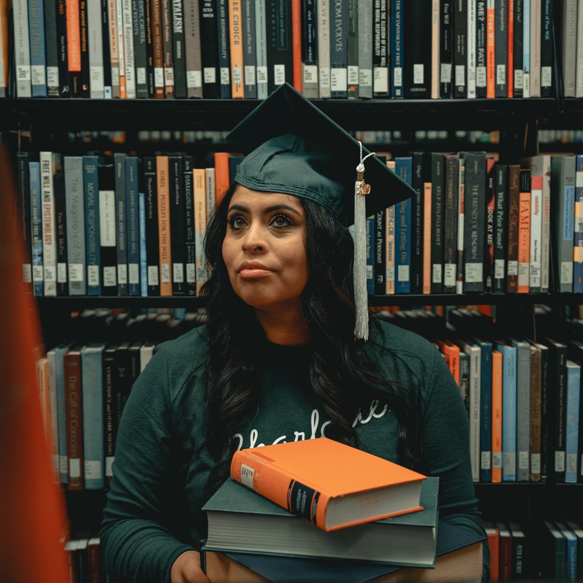 Young black student wearing graduation cap in library carrying books