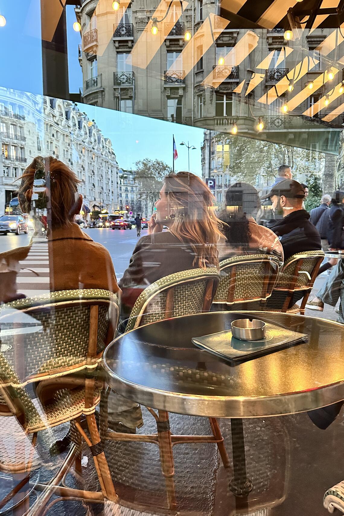 Photo is taken from inside a building, looking out through the glass. A row of chairs and small tables line the street outside; various men & women sit in the chairs facing the street as vehicles drive past in a blur.