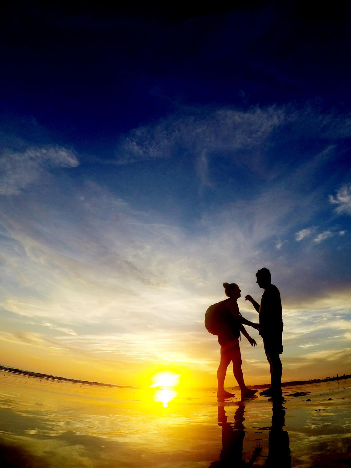 Photo is taken on a watery coast, looking up at two people standing in silhouette against a vibrant blue and yellow sunrise. The people are facing each other and gesturing; one wears a backpack and has hair tied back, the other appears to have short hair.