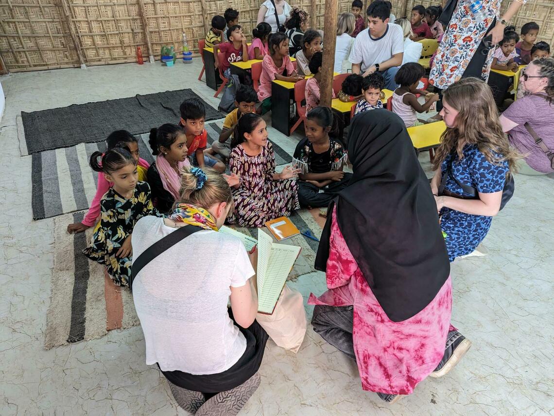 Image of a group of students in India sitting in a circle talking and taking notes with a group of local elementary aged girls.