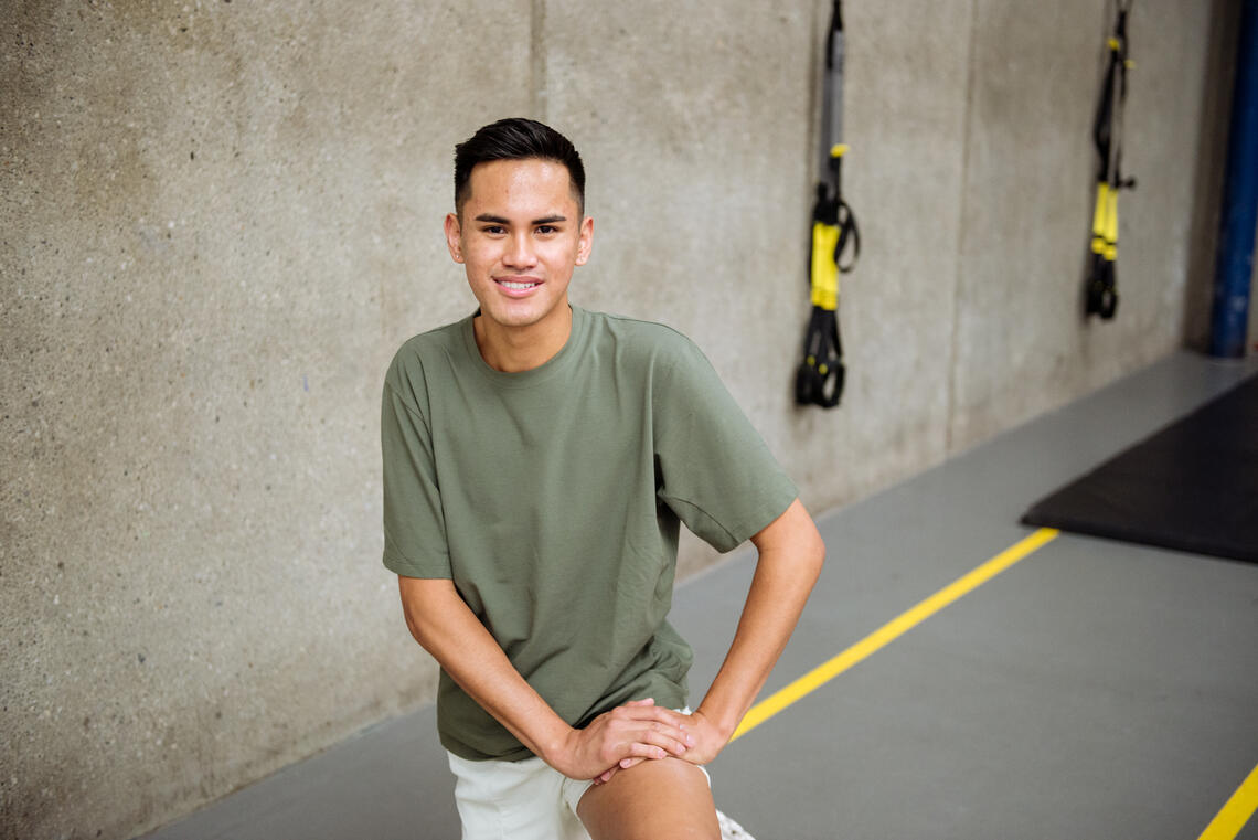 Nick Basilio, a young racialized athlete, kneels on an indoor track. He is wearing a t-shirt and shorts and smiling.