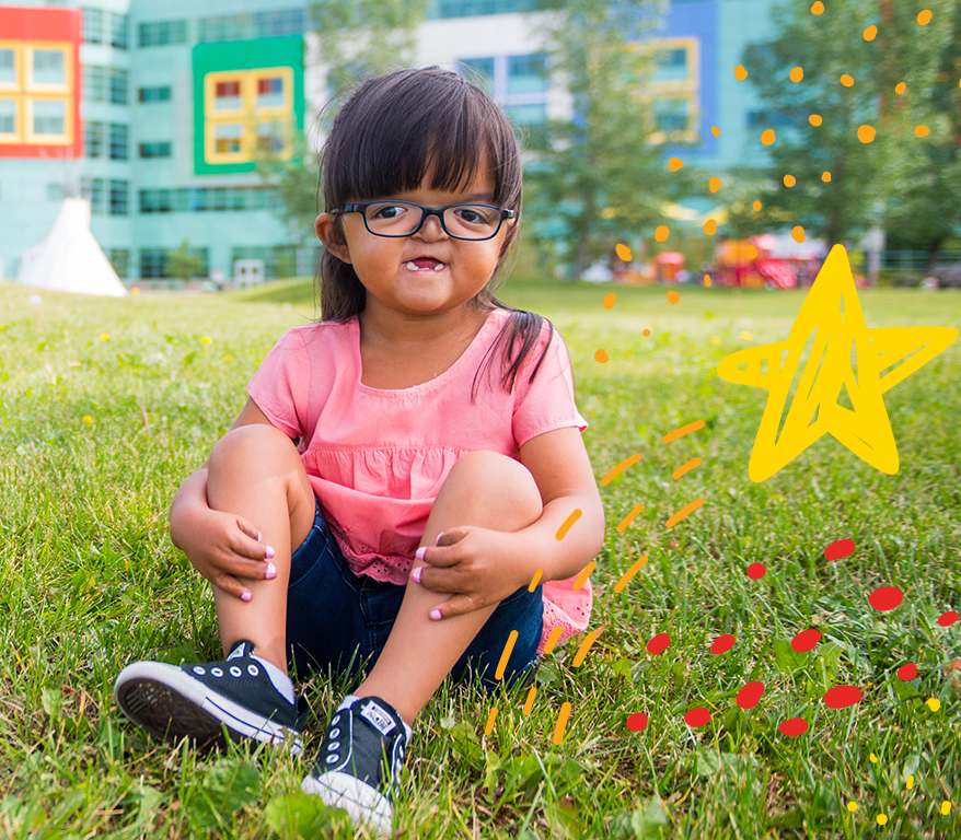 A young female child with a visible health condition sits on the grass in front of the Alberta Children's Hospital.