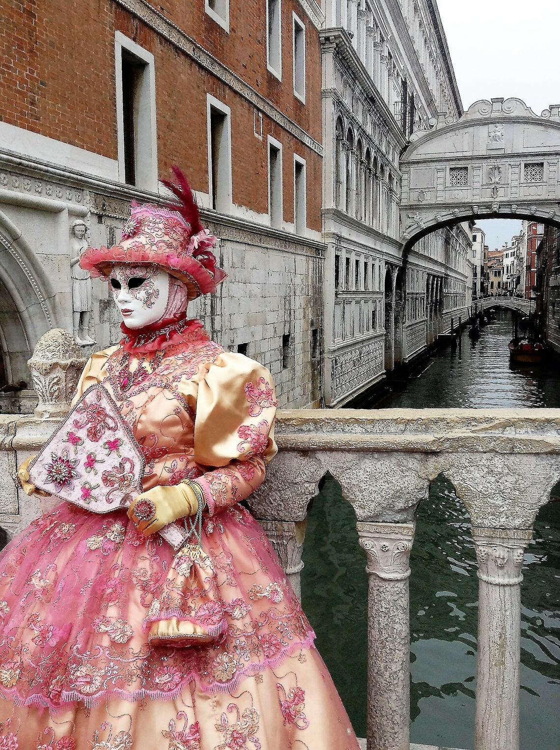 A person standing in front of a Venetian canal, dressed for carnevale in a pink and gold lace dress with a wide skirt, matching handbag and wide brimmed hat with a feather, and holding a decorative white fan. Their entire face is covered by an elaborately decorted white mask.