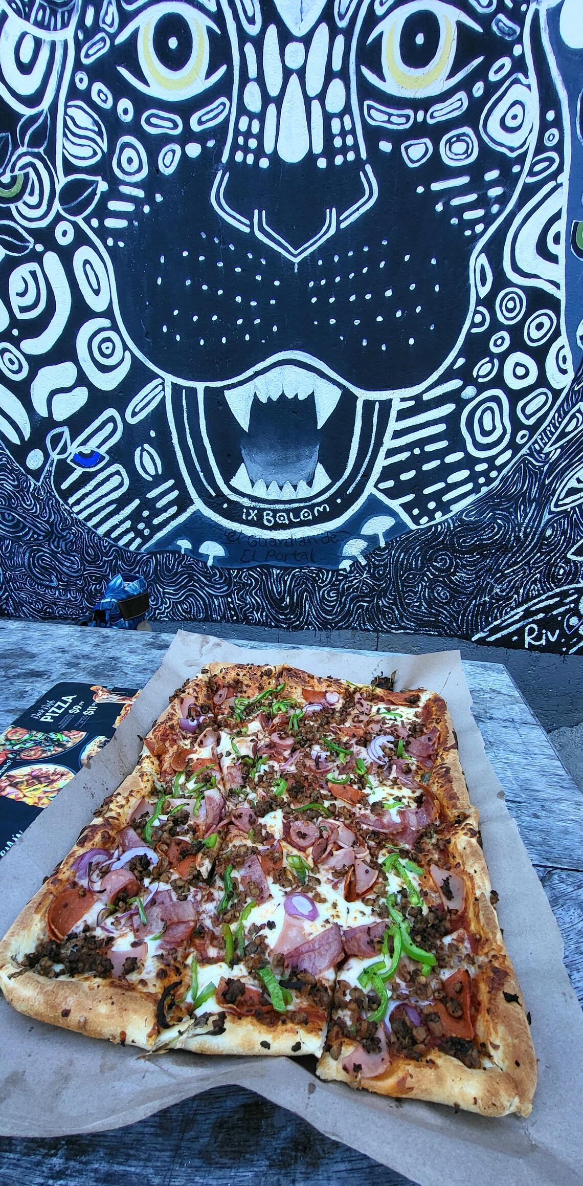 A large square pizza loaded with toppings sits on a paper sheet on a table. The wall behind the table has an artistic blue-and-white mural of a large cat with its mouth open, aligned with the pizza.
