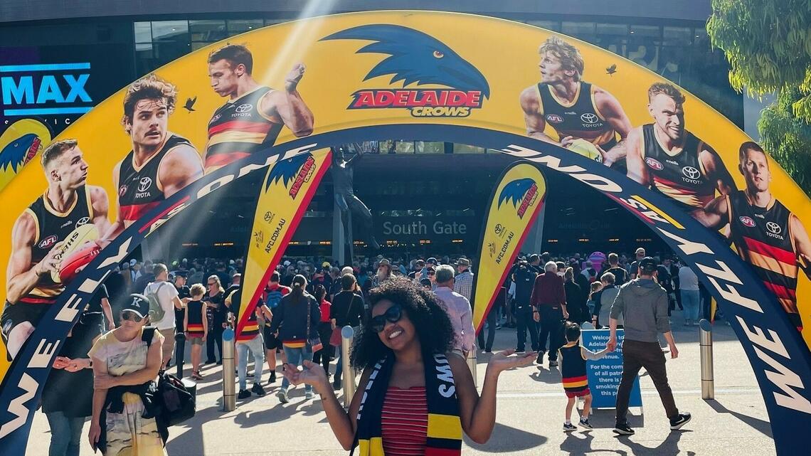 A student with dark skin and long curly hair poses in front of an Adelaide Crows decorative archway, wearing a matching scarf in the football team's colours.